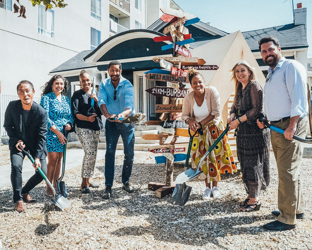 Seven people stand in front of a new home development holding shovels and smiling at the camera.