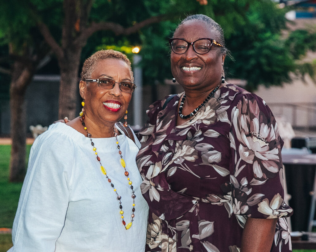 Two women of color stand side by side smiling with their arms around each other.