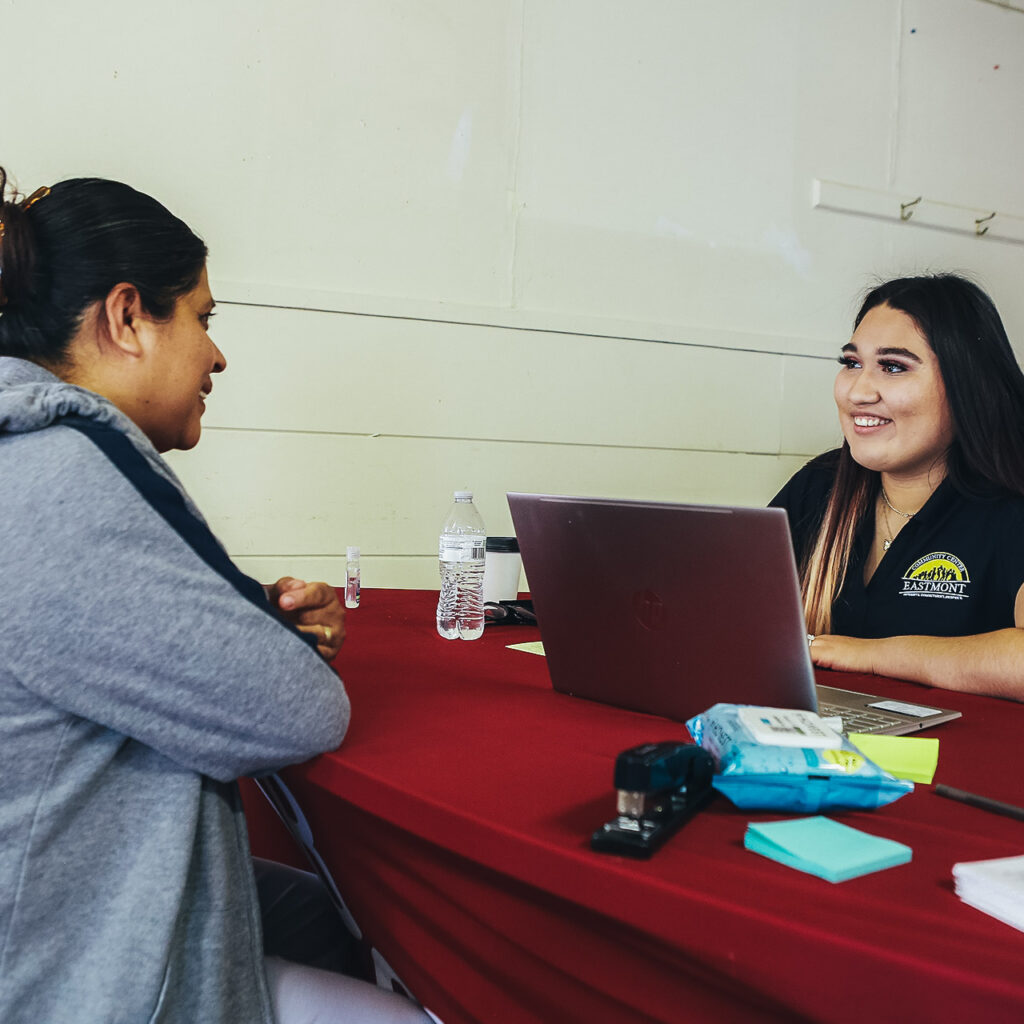 A woman at a laptop sits at a table across from another woman. They're smiling at each other.