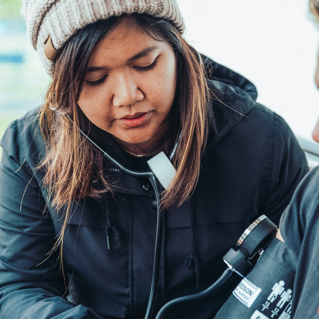 A young woman wearing a hat and coat checks the blood pressure of another person.