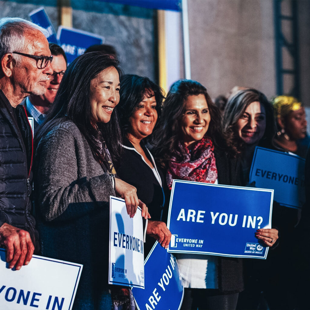 A smiling group of volunteers at an Everyone In event hold signs that read "Are you in?"