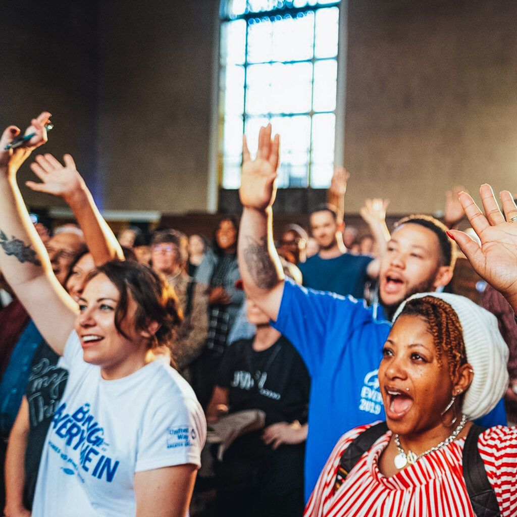 People in a crowd smile with their hands in the air as they chant something together at a rally. One woman is wearing an Everyone In LA shirt that says, "Bring Everyone In."