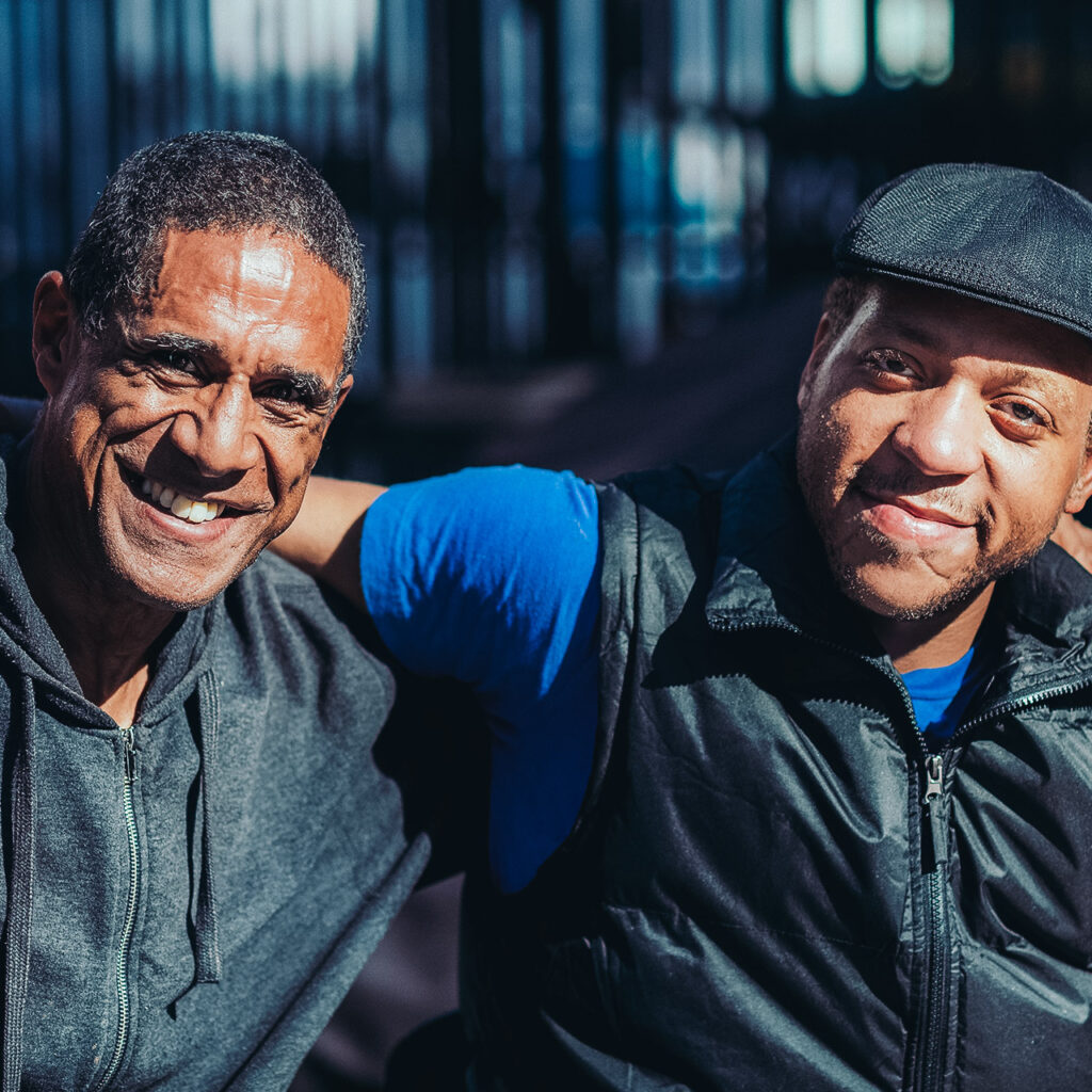 Two men smile at the camera with their arms across each other's shoulders.