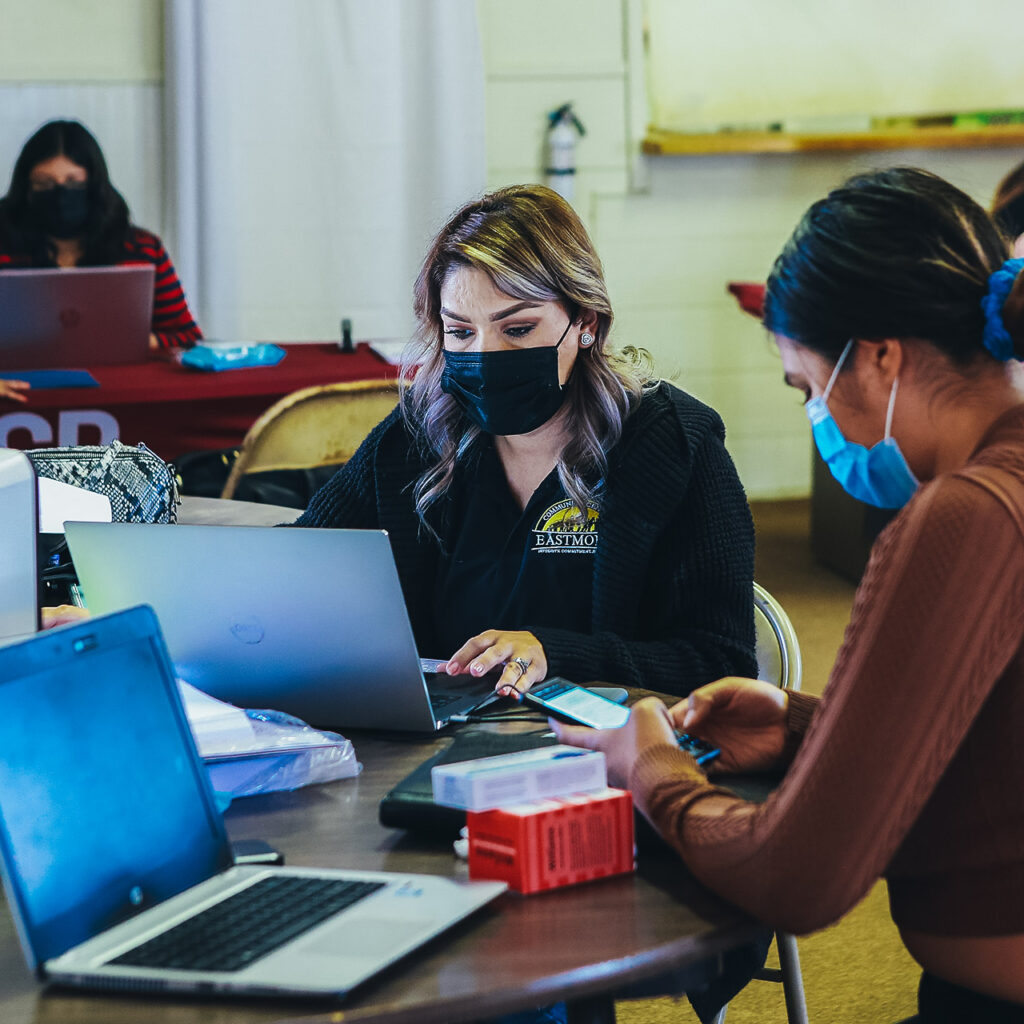 Two masked women sit at a table with laptops open