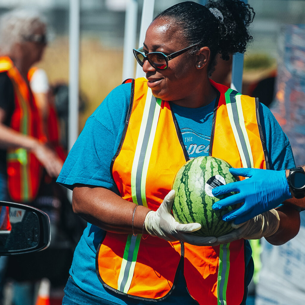 A volunteer in a bright orange safety vest carries a watermelon at a food drive.