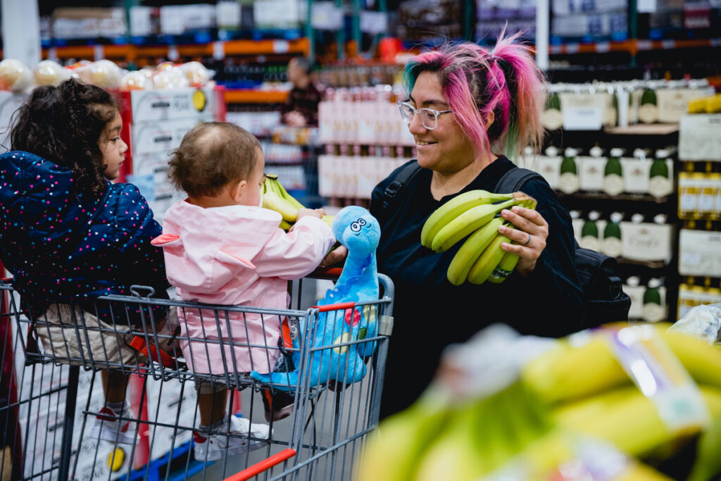 A mother in her 20s with her two young daughters are grocery shopping at a wholesale store. The kids are in a double seat shopping cart as the mom shops and pushes the cart through the store.